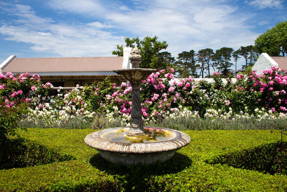 A decorative stone fountain with a classical design sits centrally in a well-maintained garden, surrounded by lush green grass and neatly trimmed hedges. The fountain features a large, circular basin at its base, filled with still water reflecting the bright sky. Above the basin, a tall pedestal supports an ornate bowl with water gently cascading from its edges. Behind the fountain, an abundance of flowering shrubs with pink and white blossoms creates a vibrant backdrop, contrasting with the rich green foliage. In the background, a building with a pitched roof and a row of tall, slender trees with dark green leaves under a partly cloudy sky evoke a peaceful outdoor setting. The garden displays elements of careful landscape maintenance typical of professional gardening work, aligning with the theme of garden care and quality landscaping offered by Gardeners Brent, as highlighted on their website.