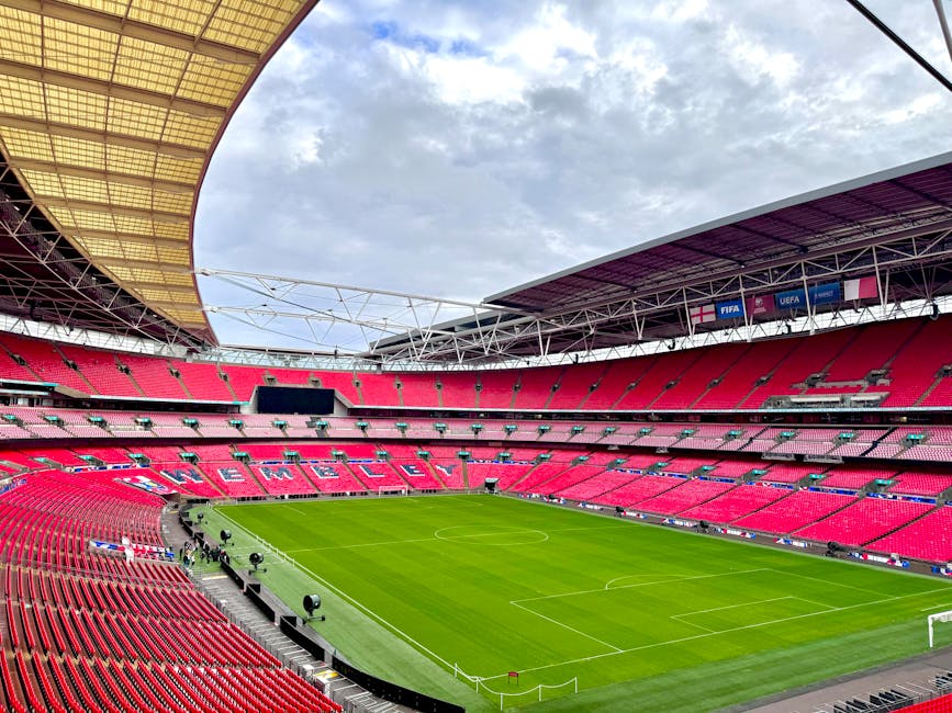 Early morning view of Wembley Stadium with a well-maintained, lush green grass pitch surrounded by rows of red stadium seats. The grass surface appears freshly mown, with visible mowing lines running across the field. The playing area is equipped with white boundary lines and goalposts at each end, while the sideline areas are bordered by a concrete pathway. The stadium's partially open roof reveals a partly cloudy sky, and the structural steel framework and banners displaying UEFA and FIFA logos are visible in the upper sections. Gardeners Brent specializes in outdoor maintenance, but in this image, the focus is on the clean, manicured lawn and the detailed layout of the stadium environment, emphasizing the importance of precise lawn care and landscaping work in large-scale sporting venues.