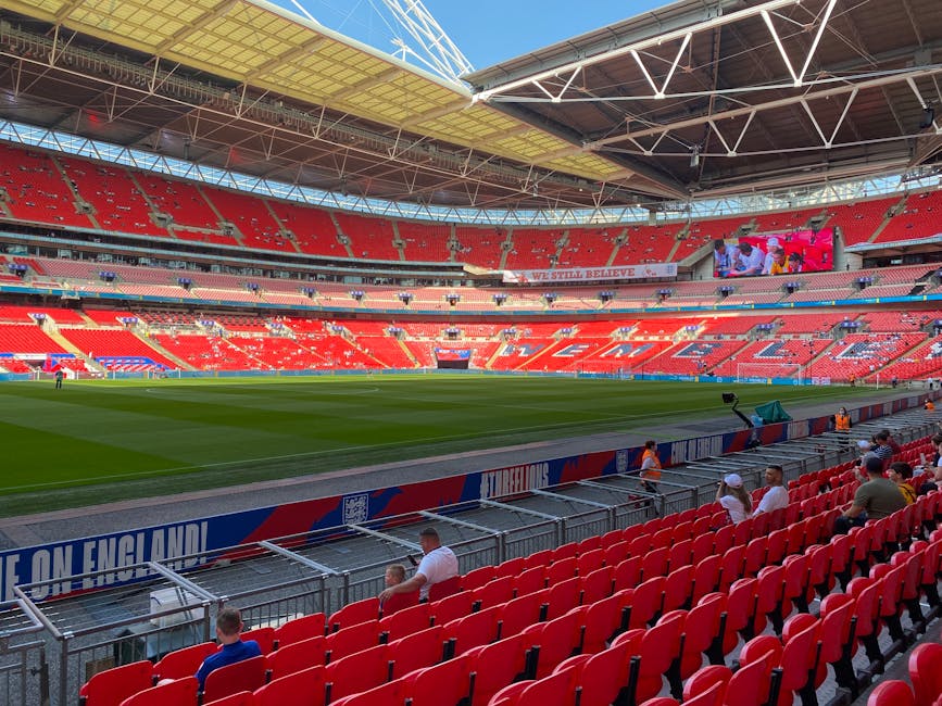 Close-up view of finely trimmed, vibrant green grass on a sports field at Wembley Stadium, with a flat perspective emphasizing the detailed texture of the blades of grass. In the background, blurred stadium seating and structural elements can be seen, highlighting the outdoor environment under clear blue skies. The scene reflects garden maintenance work, involving lawn care and turf management carried out by Gardeners Brent, with an emphasis on precise mowing and landscaping in a large sporting venue setting.